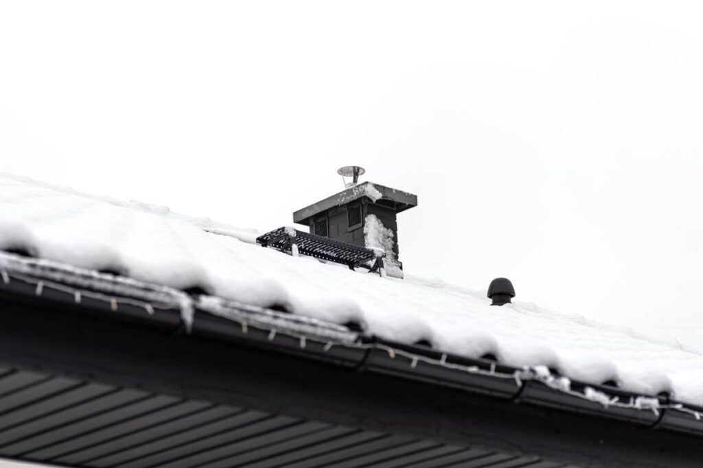 The roof of a single-family house is covered with snow against a cloudy sky, visible ceramic ventilation fireplace on the roof and falling snow.